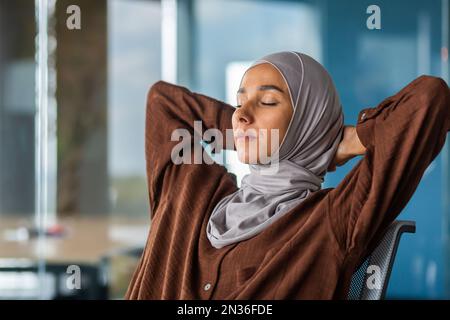 Close-up photo. Relaxed young businesswoman in hijab sitting in the office. She threw her hands behind her head, closed her eyes, leaned on a chair. She rests, meditates, dozes. Stock Photo