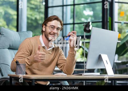 A young man poses for the camera, advertises a medicine for asthma and allergies. He sits in the office at a table with a computer, holds an inhaler in his hand, shows super with his hand, smiles. Stock Photo