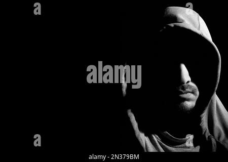 Black and white horizontal portrait shot of mysterious young bearded hooded man with his face covered in shadows isolated on dark black background wit Stock Photo