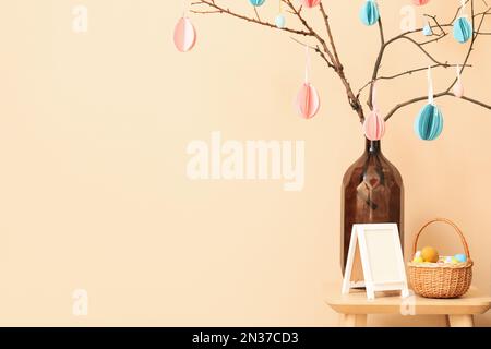 Vase with paper Easter eggs, frame and basket on table near beige wall Stock Photo