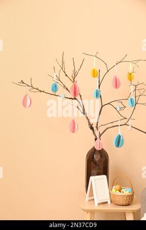 Vase with paper Easter eggs, frame and basket on table near beige wall Stock Photo