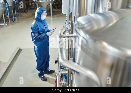 Side view at female worker wearing protective suit standing by steel tanks at industrial chemical factory Stock Photo