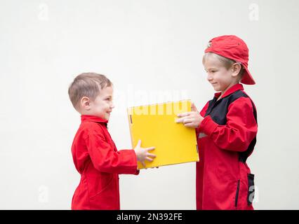 Two cute boys in red uniform stand opposite each other, hold yellow cardboard box on light background. Delivery, little postman. Work like dad. positi Stock Photo