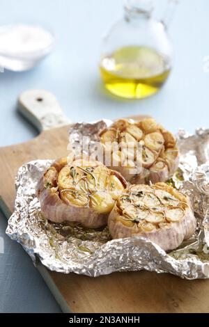 Roasted garlic bulbs with herbs and olive oil in tin foil on a cutting board Stock Photo
