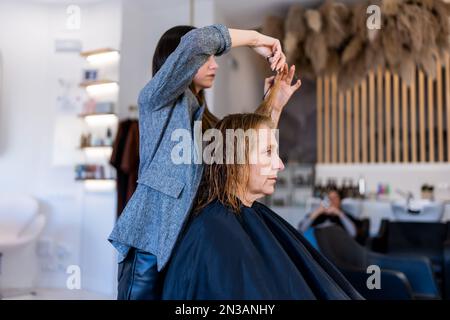 Side view of professional hairstylist cutting hair with scissors while preparing senior client for hairstyle in hairdressing salon Stock Photo