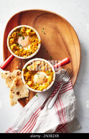 Two ramekins with lentil curry and baked eggs with naan bread on a wooden serving platter Stock Photo