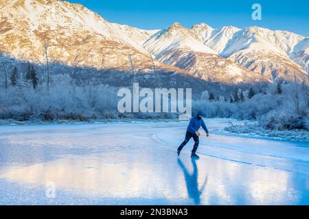 A caucasian man backcountry ice skating, nordic blading, in winter, on wild ice of Jim Creek, with the sunlit Chugach mountains behind him, and fro... Stock Photo