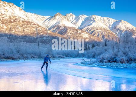 A caucasian man backcountry ice skating, nordic blading, in winter, on wild ice of Jim Creek, with the sunlit Chugach mountains behind him, and fro... Stock Photo