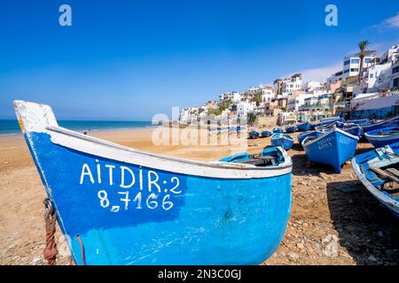 Traditional, bright blue fishing boats line the beach at Taghazout Village with the whitewashed buildings on the hillside in the background Stock Photo