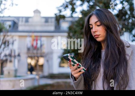 Thoughtful young female in warm coat with long dark hair browsing mobile phone while standing on city street in evening Stock Photo