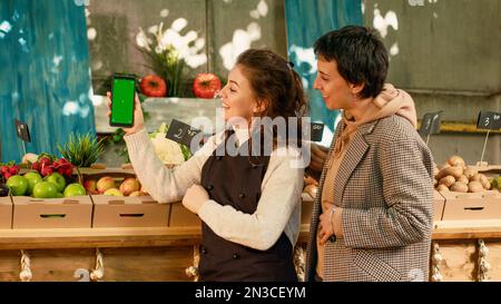 Two women looking at greenscreen display on smartphone, checking chroma key template on mobile phone app. Client and local farmer using isolated mockup copyspace template. Tripod shot. Stock Photo