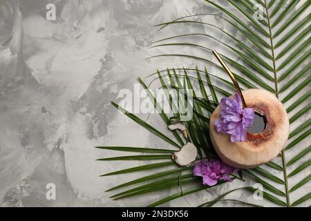 Young peeled coconut with straw, tropical leaf and flowers on grey table, flat lay. Space for text Stock Photo