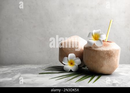 Young peeled coconuts with straw, palm leaf and beautiful flowers on grey table. Space for text Stock Photo