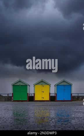 Three brightly colored beach huts sit beneath a storm cloud on a wet autumnal day; Hove, East Sussex, England, United Kingdom Stock Photo