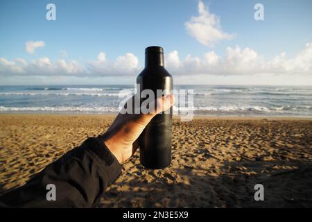 Close-up of male hand holding black thermo bottle on ocean beach at sunrise. Plastic free. Stock Photo