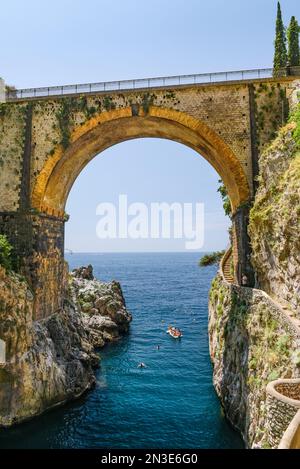 People swimming and boating under an old road bridge across the bay in Furore; Furore, Salerno, Italy Stock Photo