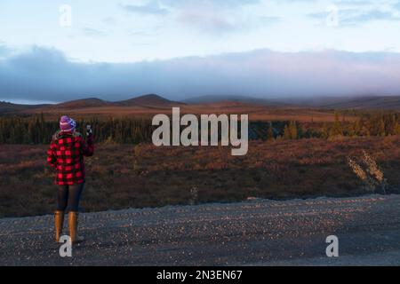 Woman taking photos with her cell phone along the Dempster Highway and enjoying the scenery in Canada's Yukon; Dawson City, Yukon, Canada Stock Photo