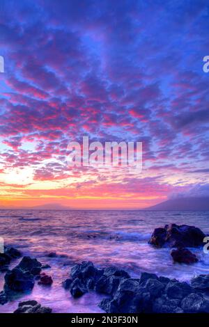 Waves wash over the lava rocks along the shore with a stunning pink and purple sunset over the Pacific Ocean Stock Photo