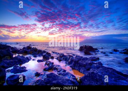 Waves wash over the lava rocks along the shore with a stunning pink and purple sunset over the Pacific Ocean Stock Photo