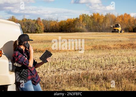 A woman standing beside a grain hauler, using portable wireless devices to manage and monitor a fall Canola harvest, talking on the phone, with a c... Stock Photo