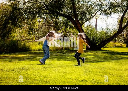 Two sisters enjoying each other's company and playing and spinning around together on a warm fall day in a city park; St. Albert, Alberta, Canada Stock Photo