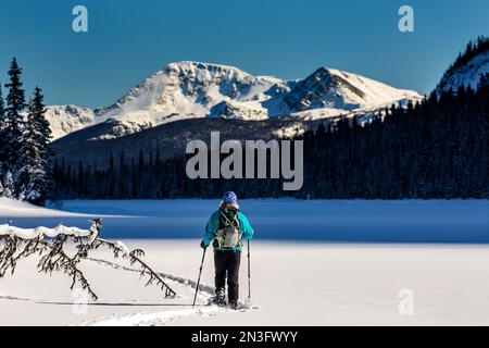 Female snowshoeing on a snow-covered lake with snow-covered Rocky Mountains in the distance and blue sky in Banff National Park Stock Photo