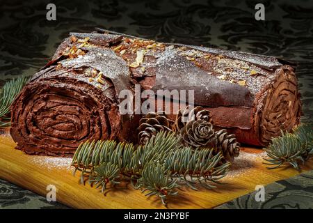 Icing frosted rolled chocolate Yule log cake with evergreen tree pieces and pine cones on a wooden board sitting on a dark green decorative table c... Stock Photo