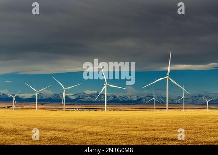 Large metal wind turbines in a stubble field with a Chinook arc, blue sky and mountain range in the distance, North of Glenwood, Alberta Stock Photo