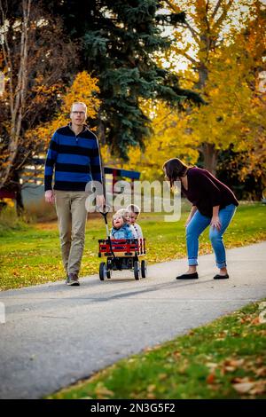 A father and mother pulling their young children in a wagon in a city park during the fall season, and their baby girl has Down Syndrome Stock Photo