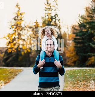 A father carrying his young daughter on his shoulders while walking in a city park during the fall season; St. Albert, Alberta, Canada Stock Photo