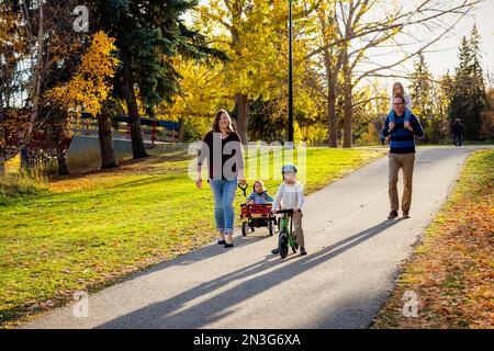 A mother pulling her daughter with Down Syndrome in a wagon, the father has their daughter on his shoulders and their son is riding his bike in a c... Stock Photo