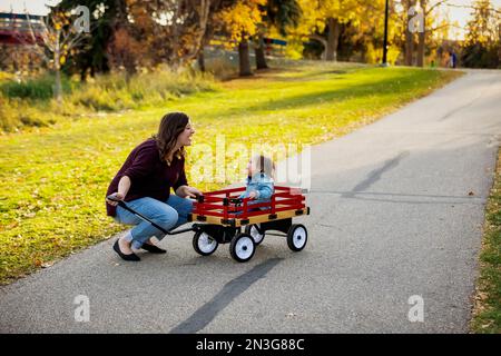 A mother pulling her baby with Down Syndrome in a wagon along a river in a city park with a bridge in the background during the fall season Stock Photo
