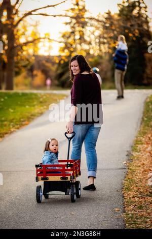 A mother pulling her baby with Down Syndrome in a wagon along a river in a city park with her family during the fall season Stock Photo