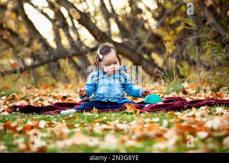 Portrait of a baby girl with Down Syndrome, sitting on the ground among the fallen leaves in a city park during the fall season Stock Photo