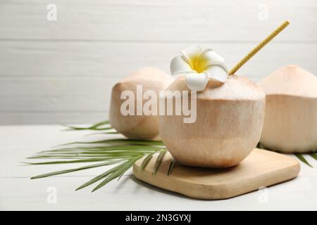 Young peeled coconuts with straw, palm leaf and flower on white wooden table. Space for text Stock Photo