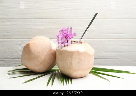 Young peeled coconuts with straw, palm leaf and flower on white wooden table Stock Photo