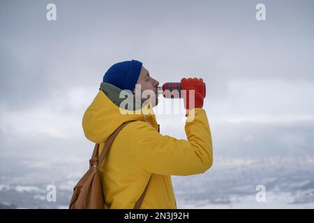 Young man tourist hiker standing on mountain top drinking hot tea from thermos during winter hiking Stock Photo