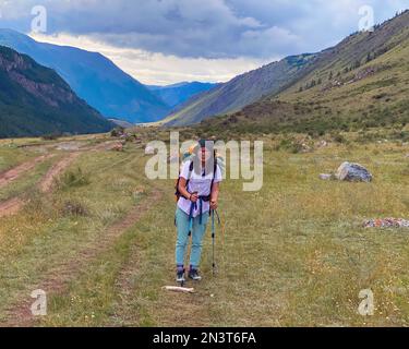 A tourist girl with a backpack and a hat stands, leaning on trekking poles, resting against the backdrop of the Altai mountains. Stock Photo