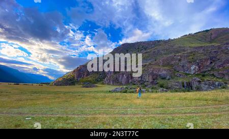 A traveler girl with a backpack walks across the field leaning on trekking poles against the backdrop of the Altai mountains. Stock Photo