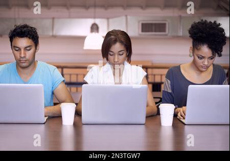 Study group dynamics. Studio shot of three students sitting at a desk working on their laptops. Stock Photo