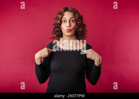 Young redhead woman wearing black ribbed dress isolated over red background pointing at herself, shocked and cheerful, self-promoting, volunteer, want Stock Photo