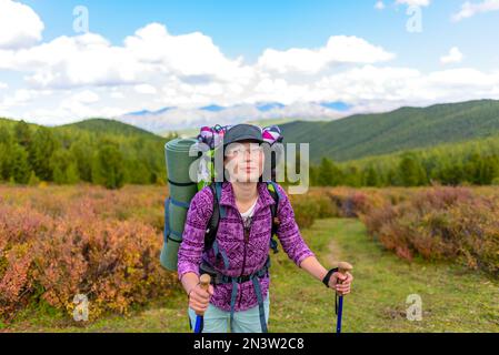 A tourist girl with a backpack in glasses and a hat stands leaning on trekking poles against the backdrop of the Altai mountains. Stock Photo