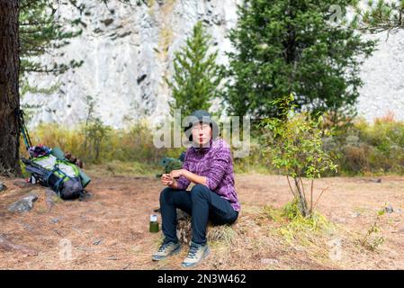 An Asian traveler girl in a hat and glasses sits on a stone near tourist things in the forest. Stock Photo