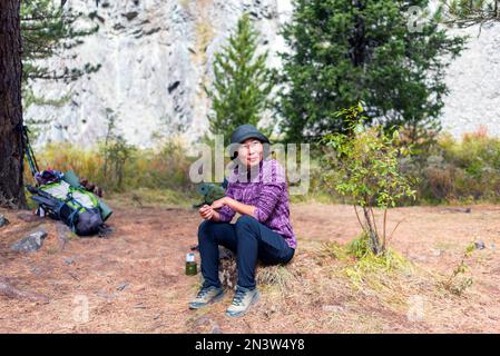 An Asian traveler girl in a hat and glasses sits smiling and relaxing on a stone near tourist things in the forest. Stock Photo