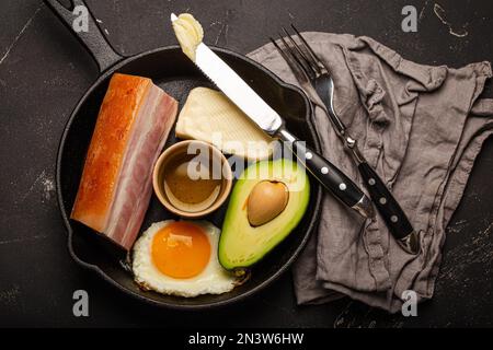 Main keto foods as butter, olive oil, fried egg, avocado, fat meat bacon for ketogenic diet on black cast iron pan on dark rustic stone background Stock Photo