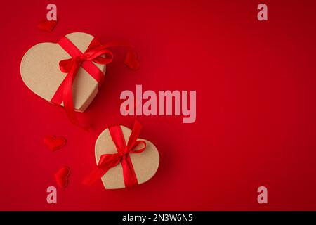 Two wrapped gift boxes in heart shape with red bow ribbon on red background and small padded hearts top view flat lay, presents for Saint Valentine Stock Photo