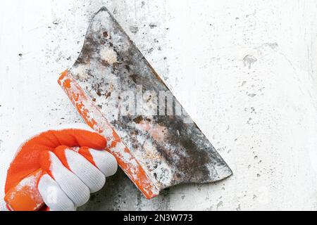 Unrecognizable hand of plasterer holds an old special putty knife and scrapes concrete interior wall, removes old plaster during redecoration of Stock Photo