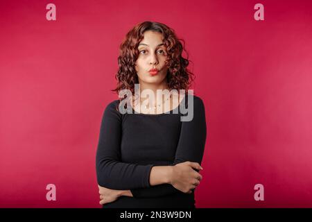 Redhead millennial woman wearing black dress standing isolated over red background shocked woman looking at the camera with open mouth. Can't believe. Stock Photo