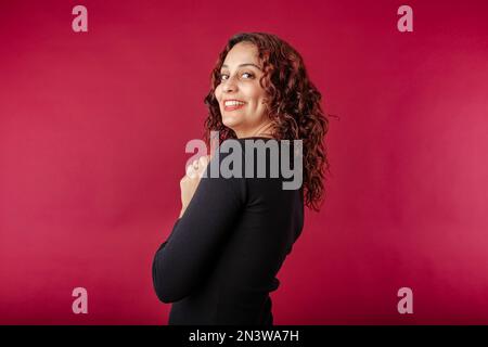 Young redhead woman wearing black ribbed dress isolated over red background looking back at the camera over shoulder with a smile. Confident and laugh Stock Photo