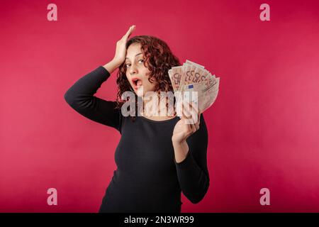 Beautiful redhead woman wearing black dress standing isolated over red background surprised looks at the camera with banknotes or money in hand and ha Stock Photo